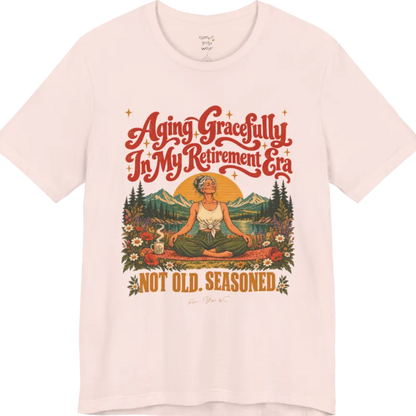 A short sleeved tee called the Seasoned T-Shirt, flat lay and a soft cotton blend shirt. Simply says, Aging Gracefully In My Retirement Era. Not Old. Seasoned. Design highlights a silver-haired figure sitting cross-legged in easy pose on a patterned mat beside a steaming mug, with a rising sun, mountains, a lake and wildflowers, and gold sparkle stars around the script lettering. Color is Soft Pink.