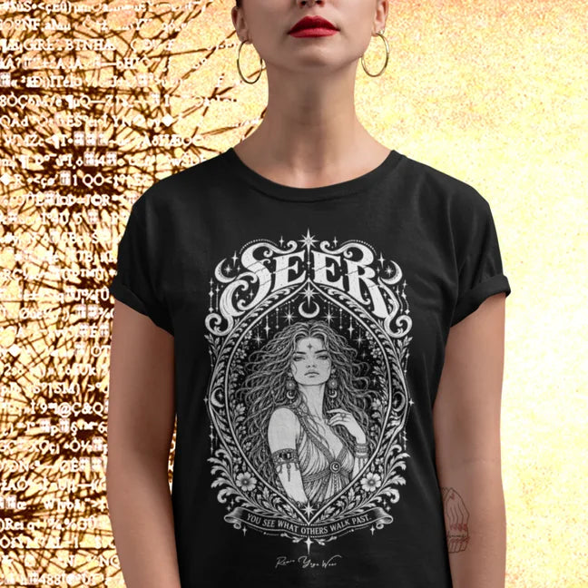A person with red lipstick and large gold hoop earrings, sleeves rolled, wearing the Seer T-Shirt with a white engraved portrait inside an ornate almond frame, standing against a warm gold background scattered with dark symbols. Color is Black.
