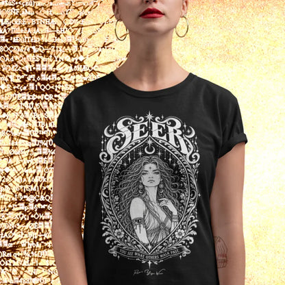 A person with red lipstick and large gold hoop earrings, sleeves rolled, wearing the Seer T-Shirt with a white engraved portrait inside an ornate almond frame, standing against a warm gold background scattered with dark symbols. Color is Black.