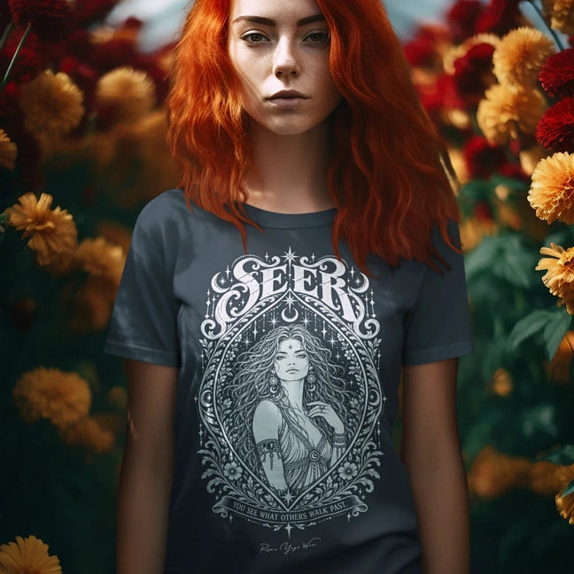 A person with long copper-red hair standing among orange marigolds and deep red dahlias, wearing the Seer T-Shirt with a white engraved portrait inside an ornate almond frame. Color is Heather Navy.