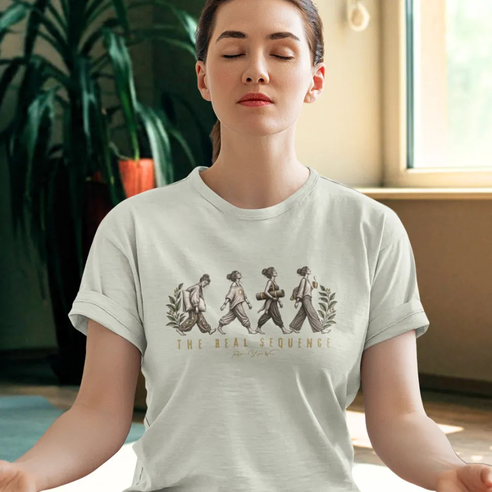 A woman stands with her eyes closed in front of a large window and houseplants, wearing the Sequence T-Shirt. Sequence T-Shirt is a Bella+Canvas 3001 and the color is Heather Prism Natural.
