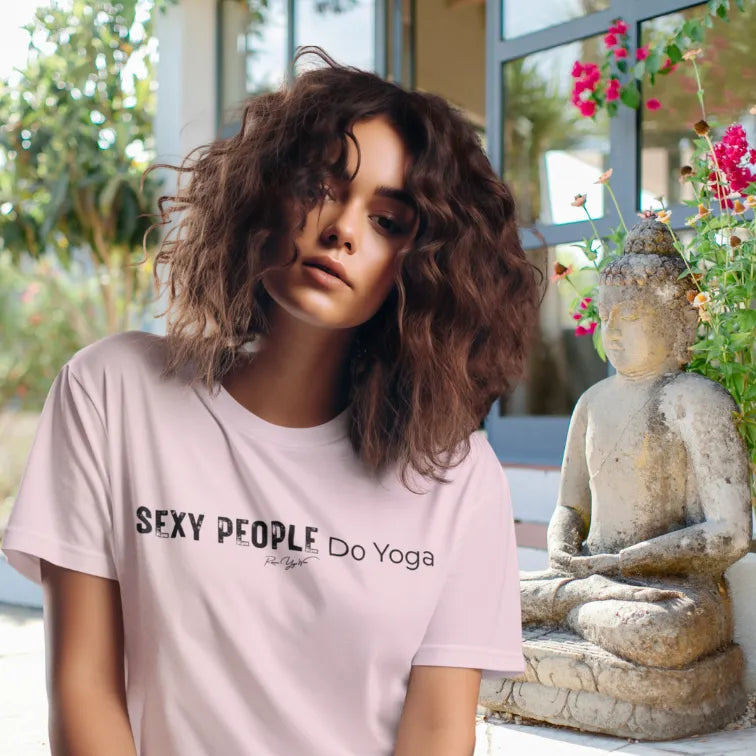 A person with curly brown hair sitting beside a stone Buddha statue in a sunny garden, wearing the Sexy T-Shirt in soft pink with Sexy People Do Yoga across the chest. Clearly ready for class.