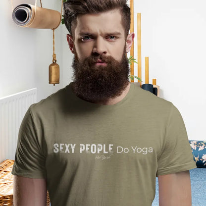 A bearded person with swept-back hair standing in a bright room with a hanging yoga mat and brass bell, wearing the Sexy T-Shirt in heather military green with Sexy People Do Yoga across the chest.