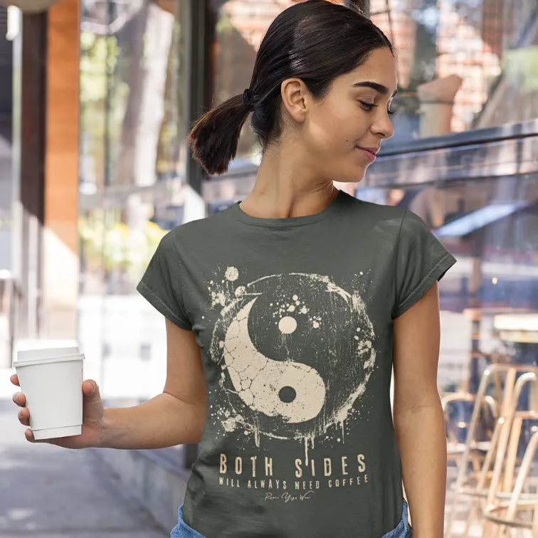 A person with a dark ponytail holding a takeaway coffee cup on a street outside a cafe, wearing the Sides T-Shirt. The tee shows a cracked grunge yin yang in cream with the words Both Sides Will Always Need Coffee. Color is heather military green.