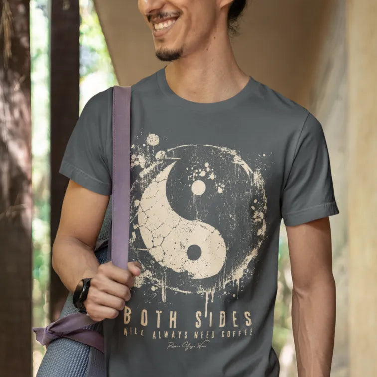 A smiling person with a goatee carrying a rolled yoga mat on a strap over one shoulder, wearing the Sides T-Shirt. The tee shows a cracked grunge yin yang in cream with the words Both Sides Will Always Need Coffee. Color is dark grey heather.