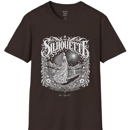 A short sleeved tee called the Silhouette T-Shirt, flat lay and a Gildan 64000 shirt on a white background. Simply says, Light Doesn't Lie About Your Shape. Design highlights a long-haired figure in a flowing gown gazing at a radiating star, framed by ornate florals, mountains and a lake, with the figure's cast shadow on the path. Color is Dark Chocolate.