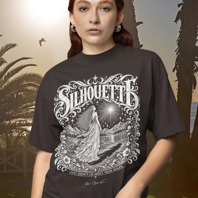 A person with auburn hair wearing the Silhouette T-Shirt in warm golden light, with palm leaves and soft shadows behind. The tee shows an ornate white linework figure gazing at a radiating star. Color is dark chocolate.