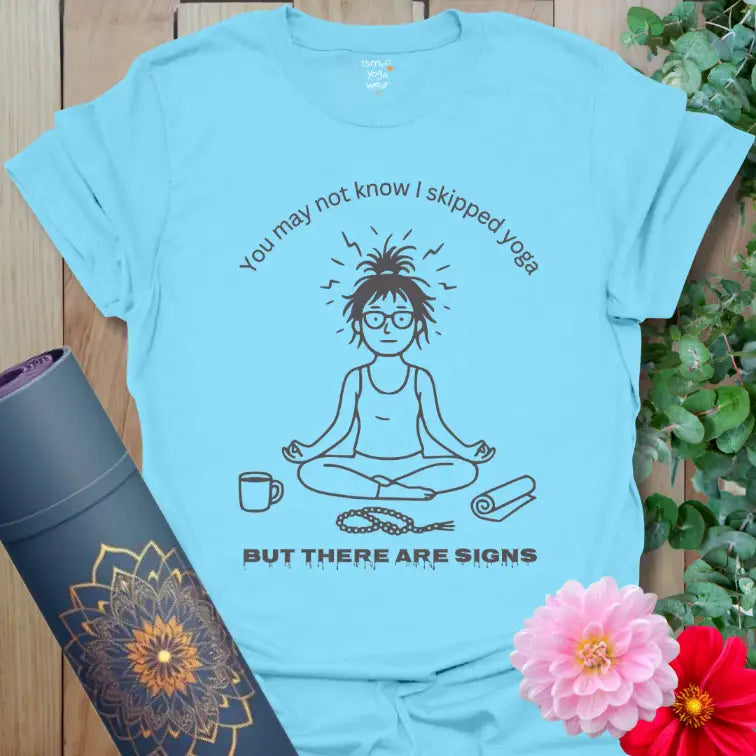 Sky blue Signs T-Shirt showing seated yogi with messy hair, coffee cup, and unrolled mat. Lighthearted yoga-lover graphic shirt.