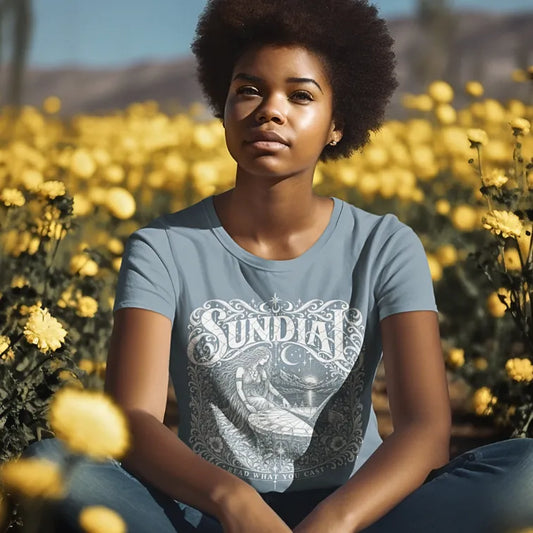 Person wearing the Sundial T-Shirt, Read What You Cast design, sitting in a field of yellow flowers, in Stone Blue.