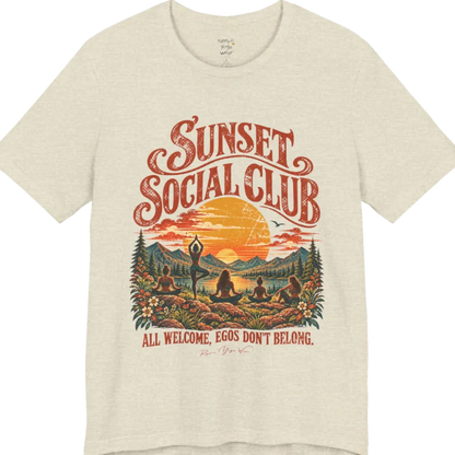 A short sleeved tee called the Sunset T-Shirt, flat lay and a Bella + Canvas 3001 shirt. Simply says, Sunset Social Club, All Welcome, Egos Don't Belong. Design highlights several people meditating together across a mountain landscape at sunset, with a large setting sun, pine trees, and florals, in a color vintage travel-poster illustration. Color is Heather Prism Natural.