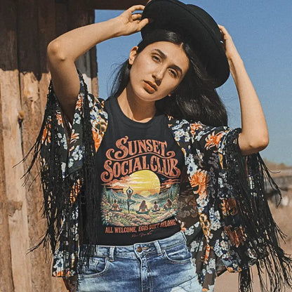 Person wearing the Sunset T-Shirt, Sunset Social Club design, outdoors against a wooden wall, in Black.