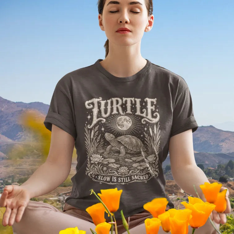 Woman wearing Turtle T-Shirt meditating outdoors among orange wildflowers, mountains behind her, shirt in Dark Chocolate.