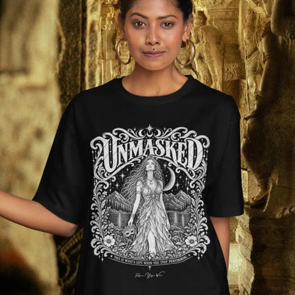 A person with long dark hair leaning against a carved stone pillar in an ancient temple, wearing the Unmasked T-Shirt in black with the Unmasked design across the chest.
