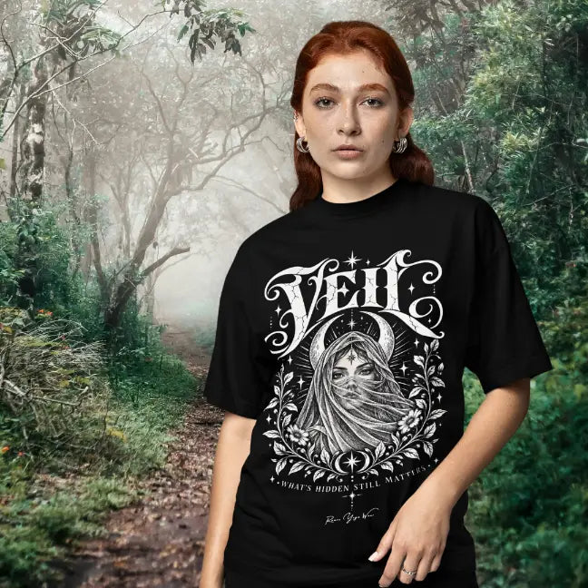 A woman stands on a misty forest path surrounded by trees, wearing the Veil T-Shirt. Veil T-Shirt is a Gildan 64000 and the color is Black.