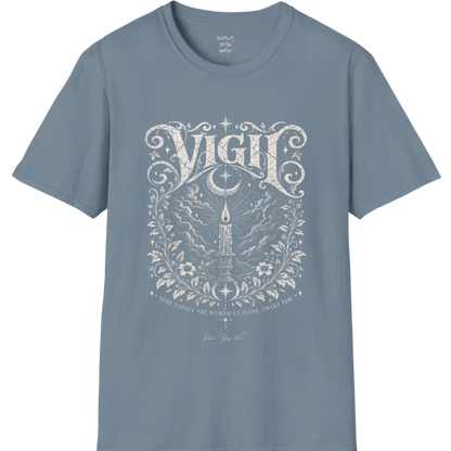 Short sleeved tee, illustration of a single candle burning steady against stormy clouds and wind lines, flame untouched, fine botanical framing at the base. Vigil T-Shirt flat lay is a Gildan 64000 and the color is Stone Blue.
