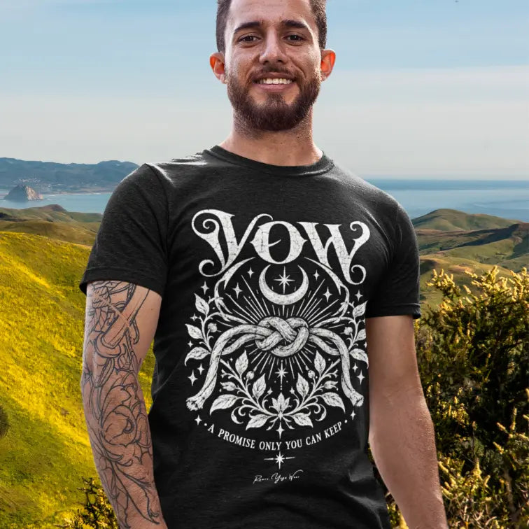 A bearded man with a tattooed arm stands on a hillside overlooking the coast, wearing the Vow T-Shirt. Vow T-Shirt is a Gildan 64000 short sleeved teed and the color is Black.