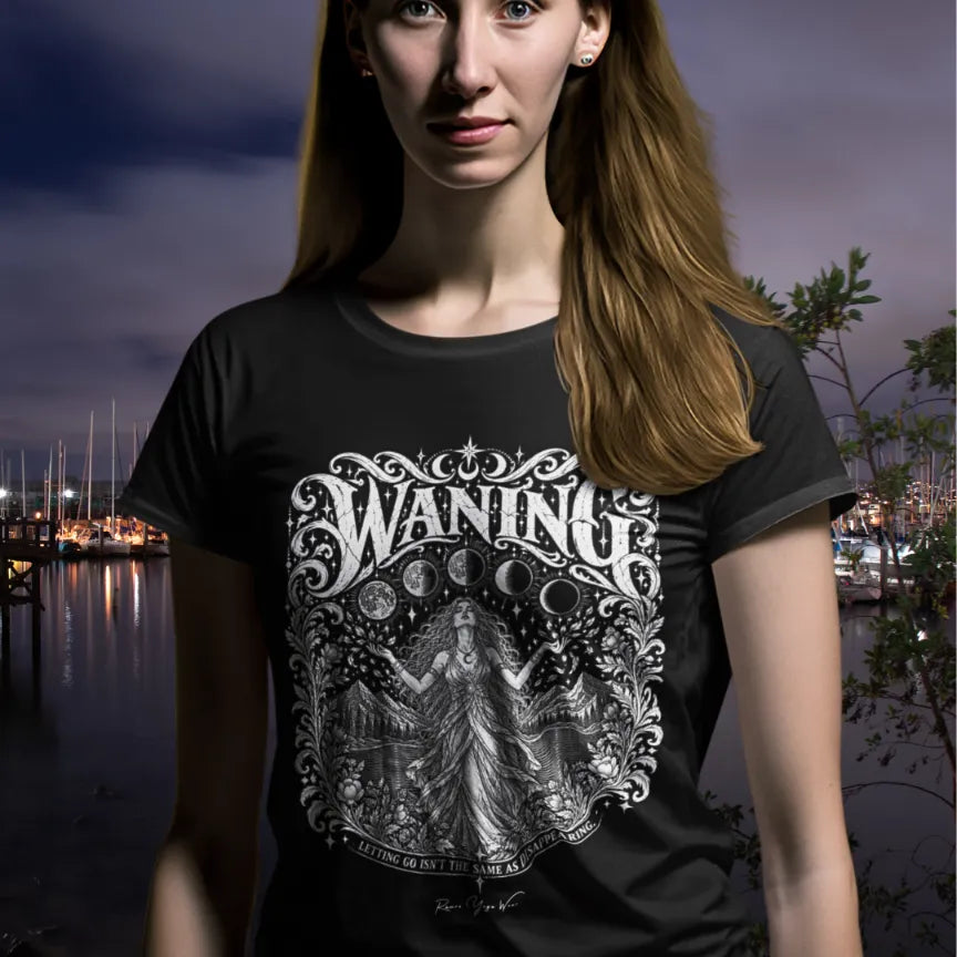 A person with long hair wearing the black Waning T-Shirt at night beside a marina with glowing lights, with the white moon-phase design printed across the chest.