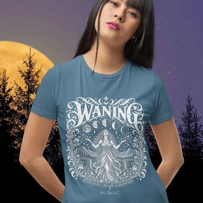 A person with dark hair and bangs wearing the Waning T-Shirt in Indigo Blue, standing in front of a large golden full moon and silhouetted trees under a starry sky, with the white moon-phase design printed across the chest.
