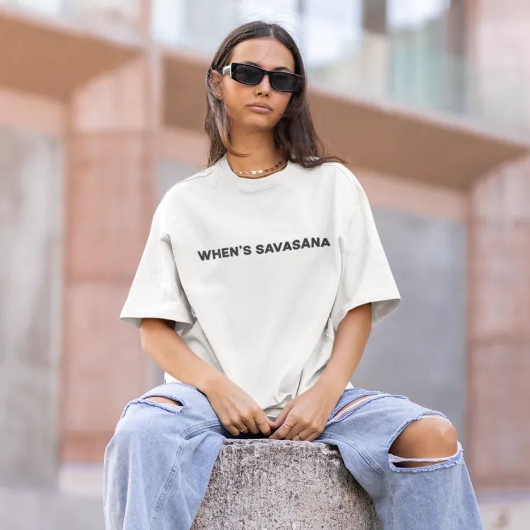 Whens Savasana T-Shirt Lifestyle 2 featuring model seated in urban setting. Clean yoga quote tee styled with relaxed fit and effortless, modern look.