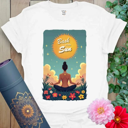 Clean white yoga t-shirt with sunrise, meditating figure, and floral elements beneath “Bask in the Sun,” made for light and intention.